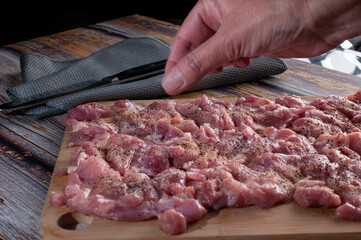 A female hand putting salt and pepper on some raw chicken cuts on a wooden cutting board with a knife on a blue napkin in the background