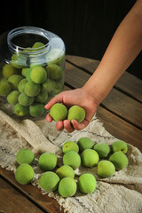 Fresh Green Plums in Hand with Glass Jar for Homemade Wine Making on Rustic Wooden Table