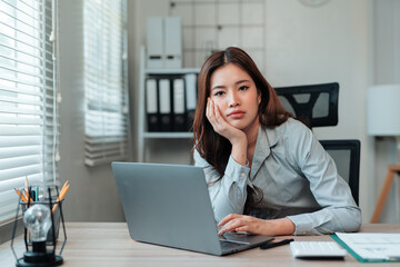 Young Asian businesswoman sitting at her desk, looking bored and tired while working on her laptop, feeling overwhelmed by workplace stress