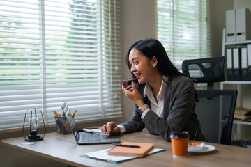 Young asian businesswoman eating a brownie while working from her office on a laptop next to a window with blinds
