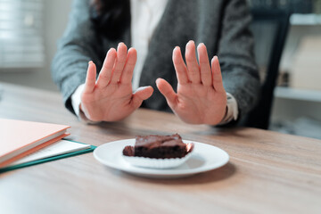 Businesswoman creating a stop gesture with her hands, firmly rejecting a tempting brownie while sitting at her office desk