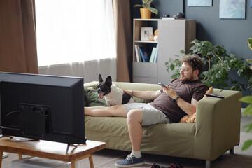 Caucasian young adult man sitting on sofa watching television holding remote control with French Bulldog beside him, relaxing in living room interior with plants and books visible
