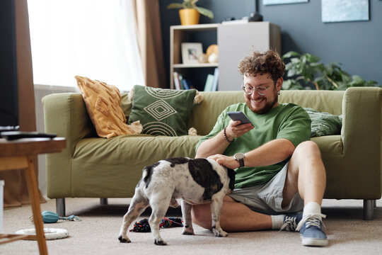 Caucasian young adult man sitting on floor smiling while using smartphone and petting small dog in living room, relaxed posture and casual interaction with pet
