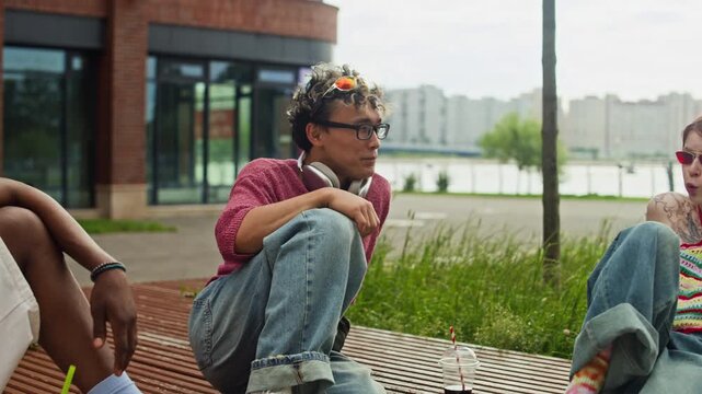 Pan shot of three diverse gen Z friends in streetwear sitting in modern city park while chatting with drinks