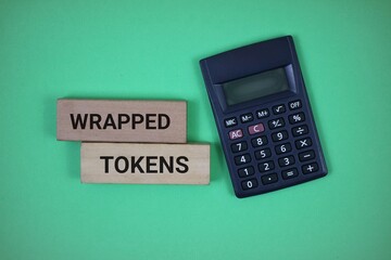 Flat lay of a calculator and wooden blocks inscribed 'Wrapped Tokens' on a green background, representing finance and modern cryptocurrency concepts.