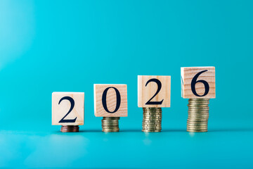 Wooden blocks with number 2026 placed on coin stacks over turquoise blue background, horizontal...