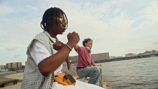 Medium long shot of intercultural male zoomers in streetwear dancing while sitting on city embankment in summer