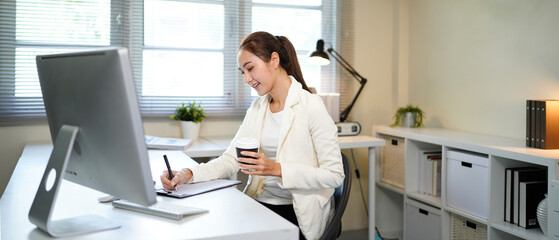 A woman is sitting at a desk with a computer monitor and a cup of coffee. She is smiling and she is...