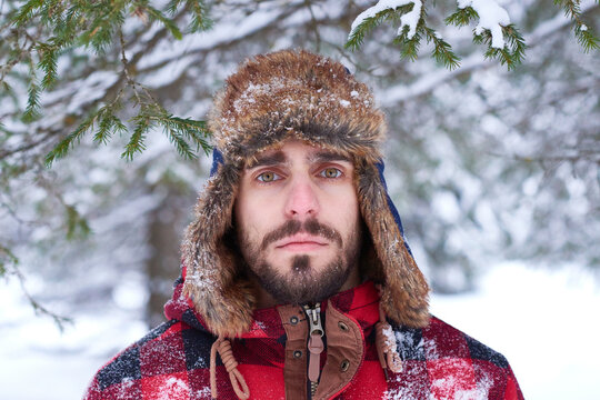 Portrait of Caucasian young adult man wearing winter hat, standing outdoors in snowy forest looking directly at camera with serious expression snow covering clothing and hat