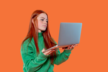 Young woman with long red hair engaged in using a laptop while wearing a green sweater against a vibrant orange background