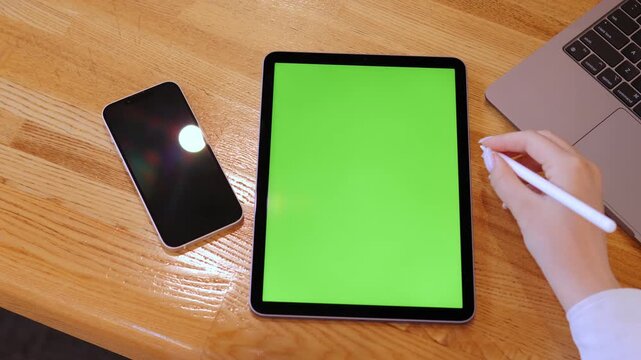 Person is writing on a green tablet with a pen. A cell phone is on the table next to the tablet