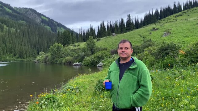 TTA man in a green warm sweatshirt drinks his morning coffee while walking along the shore of the Middle Kolsai Lake. Beautiful green area with forests and hills. Adventures in Kazakhstan.