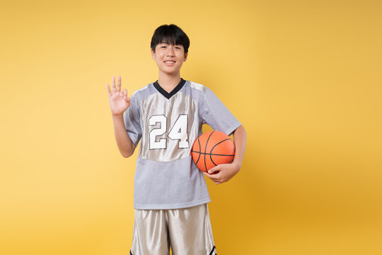 Teen basketball player in uniform holding ball and making an OK sign on yellow background - Powered by Adobe