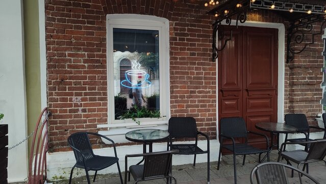 Street cafe in city. Glowing neon sign in window of brick building. Cozy coffee house terrace with glass tables and metal chairs