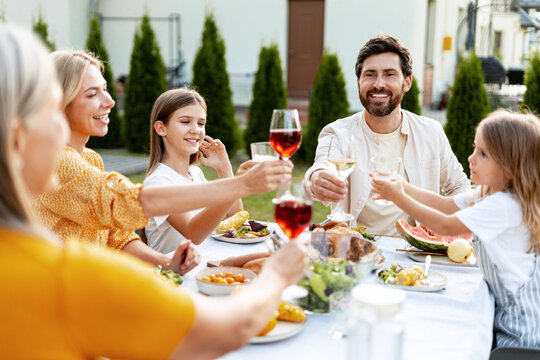Beautiful family toasting during lunch in garden at home, giving thanks sitting at festive table - Powered by Adobe