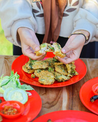 Hands breaking crispy vegetable fritters served on red plate with sambal chili dip