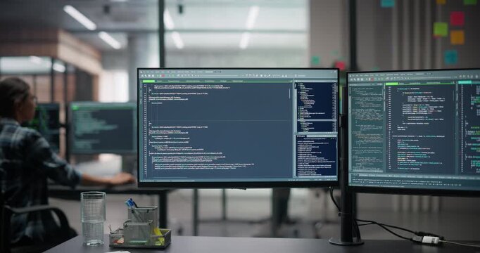 Software Developer Office Workplace with a Desk with Two Computer Screens with Programming Code, Big Data Models, Back-End of AI or Neural Network Technology Systems. Female Working in the Background