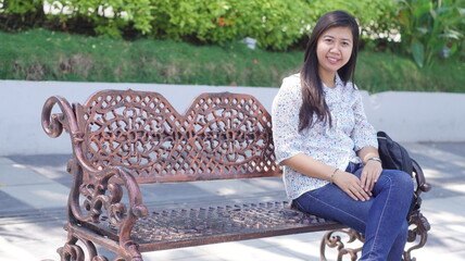 Young Asian woman sitting on a decorative bench in the park, wearing a casual floral blouse and jeans, enjoying a relaxing moment outdoors with natural greenery background