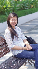 Young Asian woman sitting on a decorative bench in the park, wearing a casual floral blouse and jeans, enjoying a relaxing moment outdoors with natural greenery background
