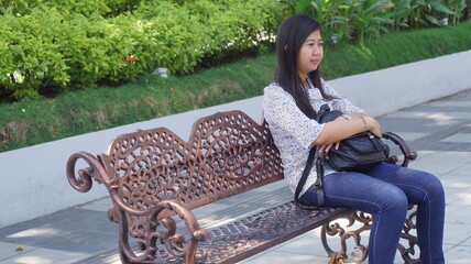 Young Asian woman sitting on a decorative bench in the park, wearing a casual floral blouse and jeans, enjoying a relaxing moment outdoors with natural greenery background