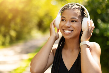 Closeup of cheerful African American woman with braided hair smiling while listening to music outdoors. Healthy lifestyle and relaxation in park
