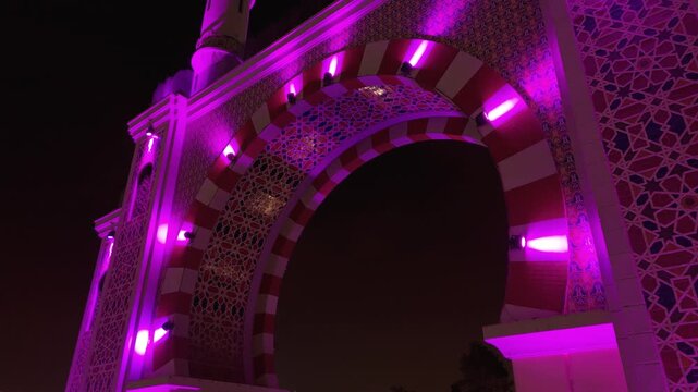 An aerial close-up shot reveals the intricate Moorish patterns and architectural details of the Arco de la Amistad, illuminated by vibrant purple lights at night in Lima, Peru