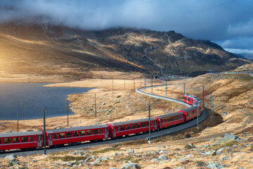 Aerial view of red Bernina Express train, curving railroad, turquoise lake in the Swiss Alps, rocky hills and dramatic cloudy sky at sunset in autumn. Alpine mountains. Top view of train in fall