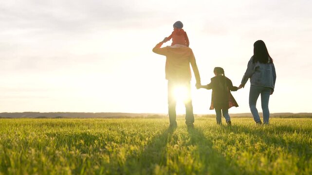 Walking through grass field with family father mother holding child hand while daughter and son explore green meadow at sunset together parent sharing joyful walk and connection in open nature
