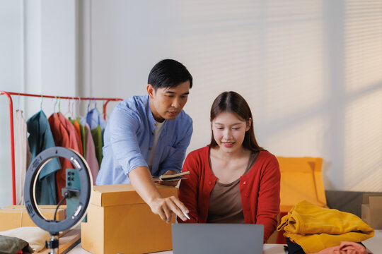 Two young asian entrepreneurs working from home are checking online orders on a laptop and preparing packages for shipping, using a ring light for better lighting