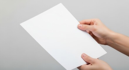 Obraz premium Two hands hold a blank white paper at an angle against a plain light gray background.