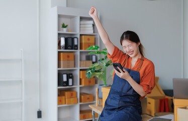 Young asian woman wearing apron raising her fist while looking at mobile phone and celebrating success in startup office, small business owner working at home office selling online on a website
