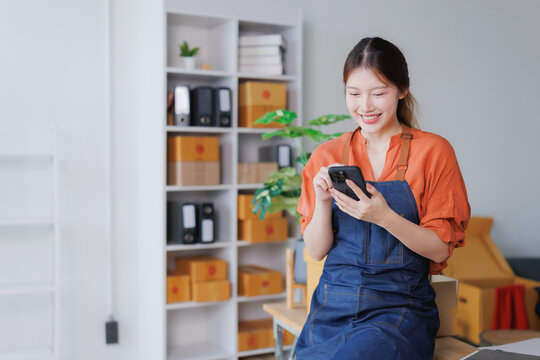 Happy young Asian female entrepreneur wearing an apron, standing in her office or storage room, smiling and using a smartphone, managing online orders and social media for her small business