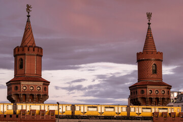 Oberbaum Bridge in Berlin with subway crossing at sunset, twin towers and arches symbolizing architectural heritage, mobility and connection over Spree River