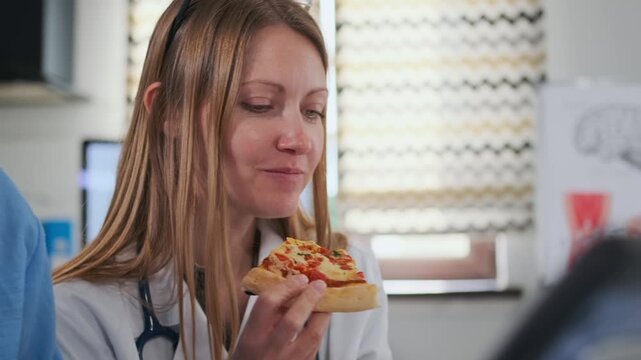 Close-up female doctor in white coat smiling and eating a slice of pizza during lunch break in bright modern medical office. Concept of work-life balance, relaxation and informal moments at workplace.