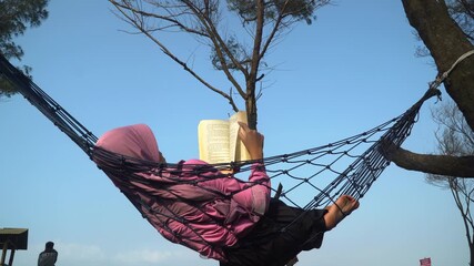 Girl wearing hijab reading a book while relaxing in a hammock