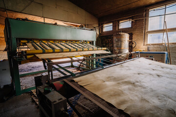 Industrial machine processing wood veneer sheets in a workshop with conveyor rollers and natural window light highlighting the manufacturing environment.