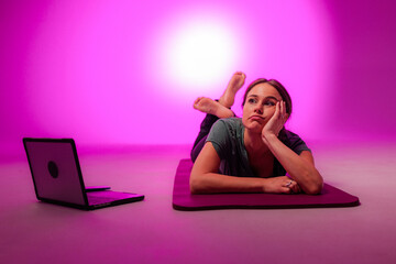 Bored woman lying on yoga mat ignoring online fitness class on laptop