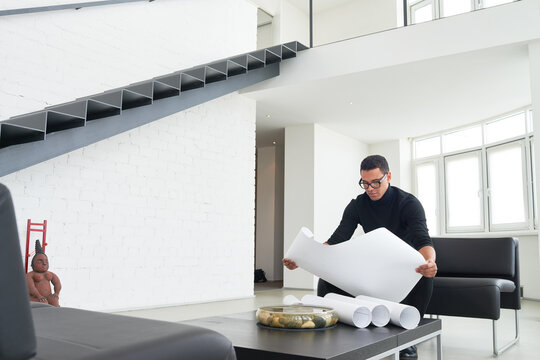 Young adult man wearing glasses examining architectural blueprints in modern office interior, rolled plans on table - Powered by Adobe