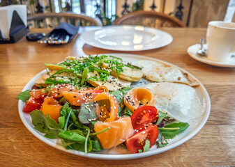 Healthy brunch bowl featuring smoked salmon mixed greens corn fritters eggs and cherry tomatoes in modern cafe setting.Gourmet salmon, vegetable salad with golden corn cakes.Artisanal breakfast dish