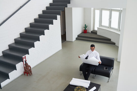 Young adult man sitting on black sofa reviewing large architectural blueprint in modern minimalist office interior, rolled papers on table nearby - Powered by Adobe