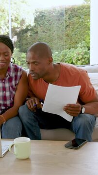 Vertical video: African American couple reading paper on sofa pointing key lines, cheering with mug