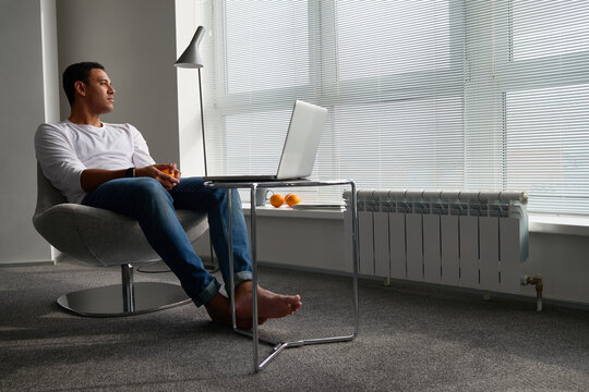 Young adult man sitting barefoot in modern chair near window, looking outside while holding hands together, laptop on glass table in front, natural daylight streaming through blinds - Powered by Adobe