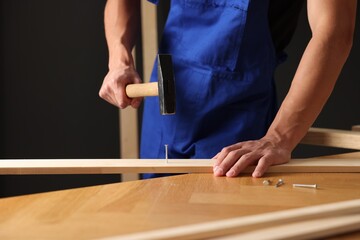 Professional repairman hammering nail into wooden plank indoors, closeup