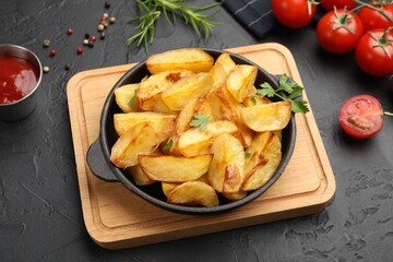 Tasty baked potatoes with parsley in frying pan, spices and tomatoes on dark textured table