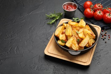 Tasty baked potatoes with parsley in frying pan, spices and tomatoes on dark textured table. Space for text