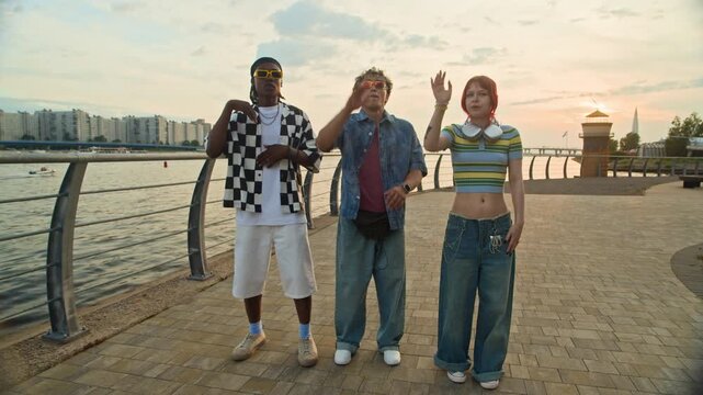 Long POV shot of three multiethnic zoomers in urbanwear practicing viral dance moves on camera while standing on city embankment, UGC