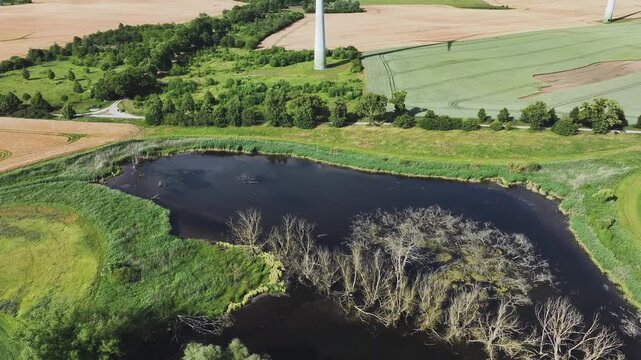 Aerial view of wind turbines standing tall over fields and a dark lake surrounded by green vegetation, a striking contrast of industry and nature, Br&uuml;ssow Uckermark, Brandenburg, Germany.