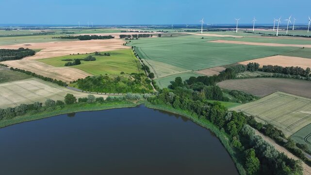 Aerial view of a serene lake surrounded by lush greenery and agricultural fields dotted with wind turbines under a clear blue sky, Br&uuml;ssow Uckermark, Brandenburg, Germany.