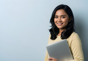 Obraz premium A young woman with dark hair and a warm smile holds a laptop against a light blue background.