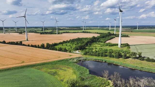 Aerial view of wind turbines standing tall in golden fields, alongside a dark, winding river and lush green vegetation, Br&uuml;ssow Uckermark, Brandenburg, Germany.